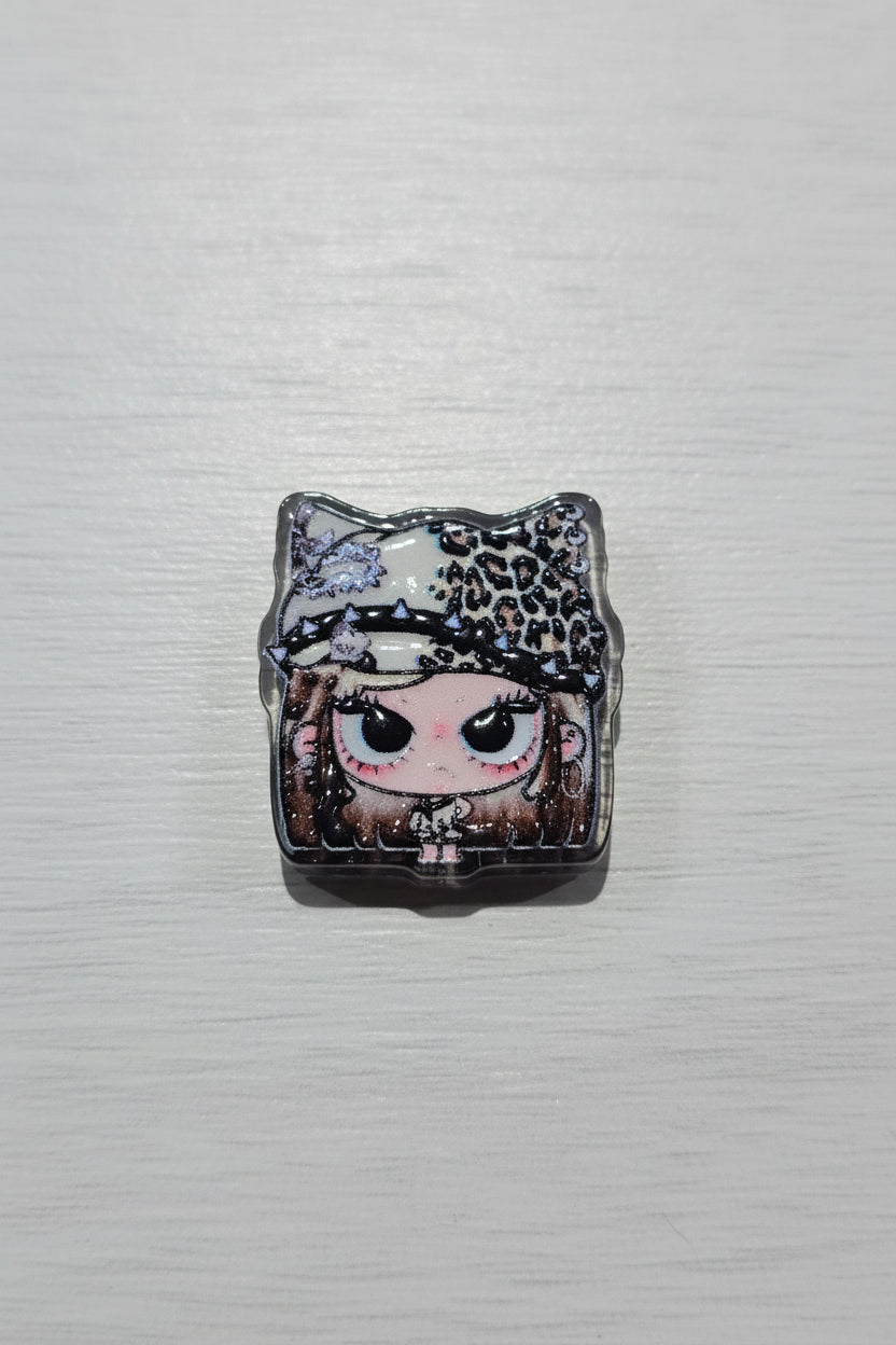 A square acrylic bead featuring a stylized anime doll face with large black eyes, a leopard-print hat, and glittery details.