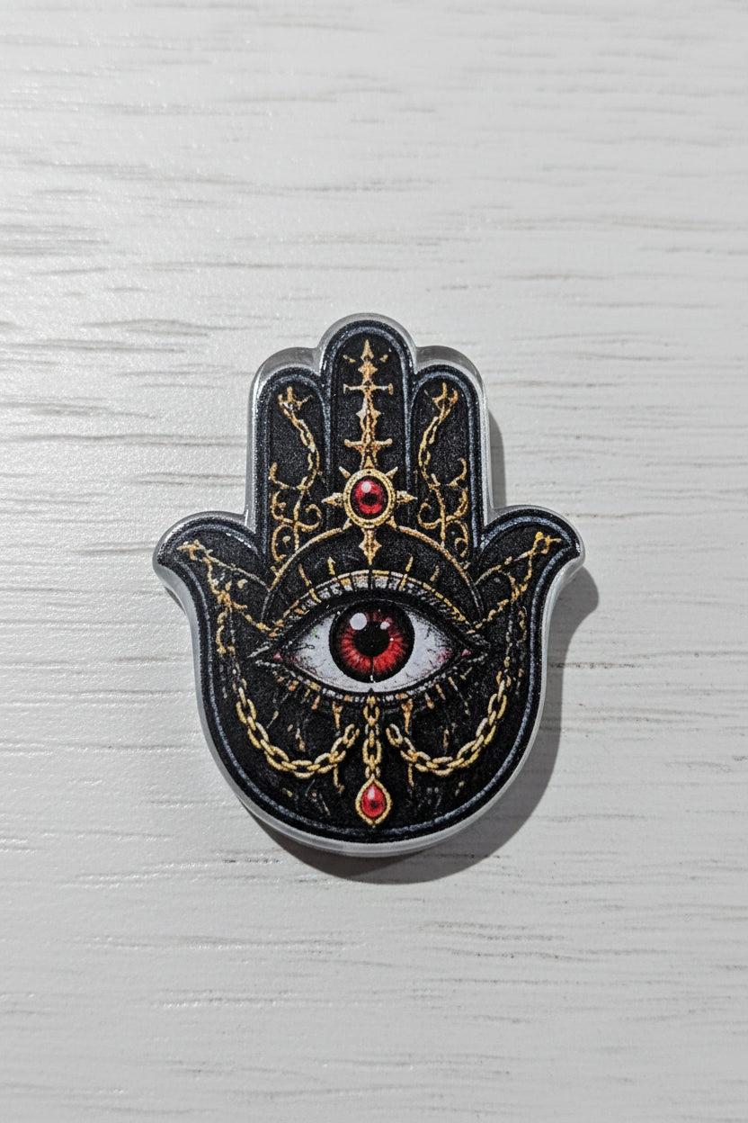 A Hamsa hand bead featuring a black and silver textured background with gold filigree patterns, a central red and white eye, and small red gemstone accents.