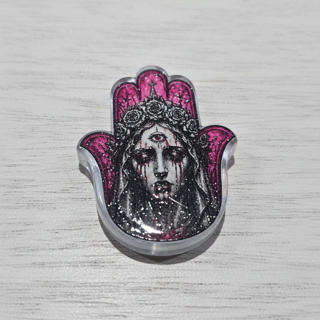A gothic-style hamsa hand bead featuring a black and white illustrated face with red blood-like streaks, a silver-toned metal border, and vibrant pink patterned accents on the fingers.