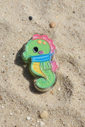 A small seahorse-shaped accessory with a green body, pink top, blue scarf, yellow belly, and black eye detail.