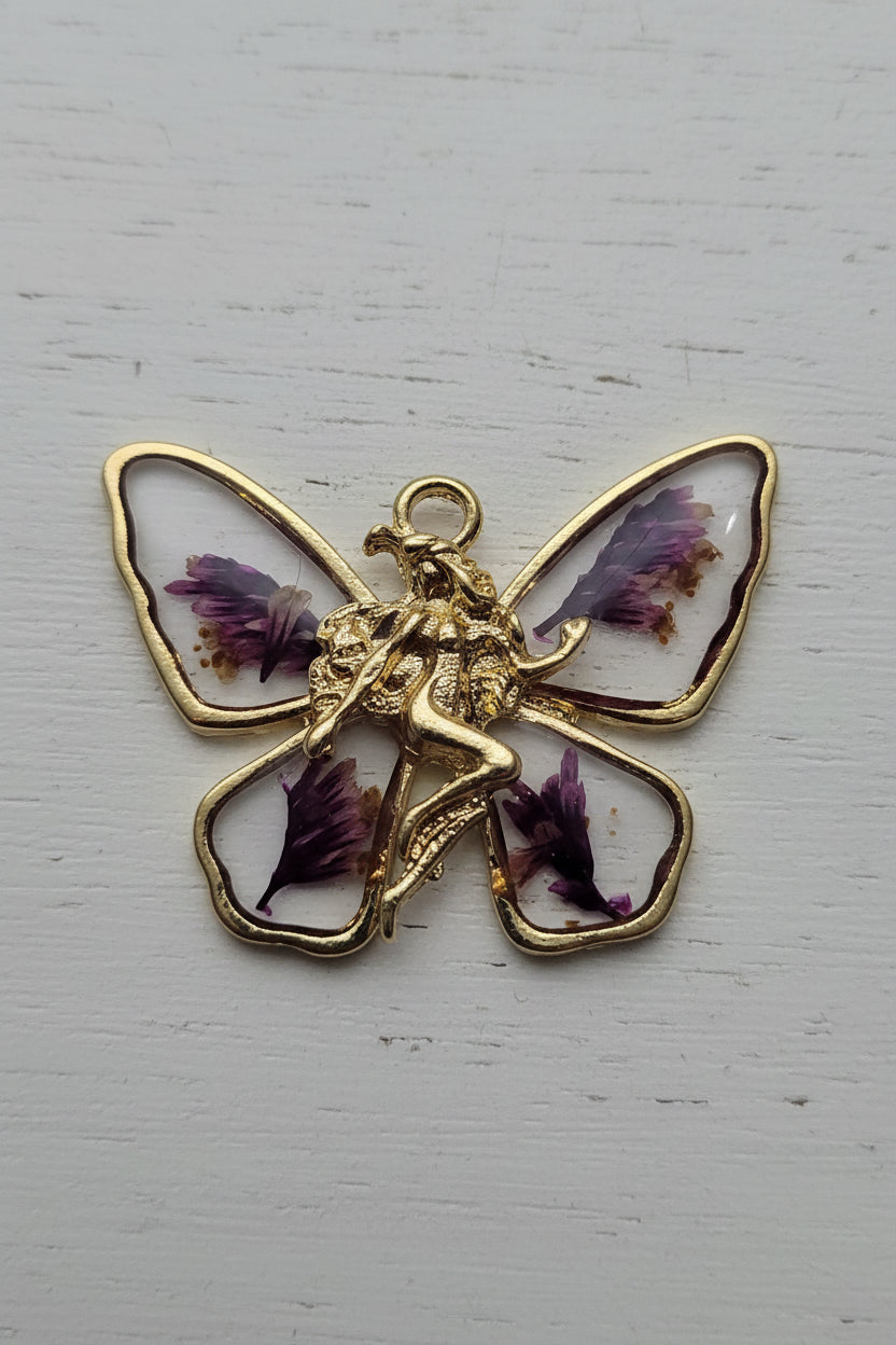 A butterfly-shaped pendant or charm with gold-tone metal outlines and clear wings containing dried purple flower petals.