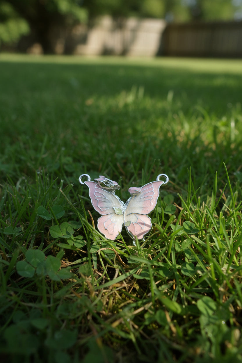 A butterfly-shaped jewellery charm with light pink enamel, silver-toned metal borders, and a small green accent near the centre.