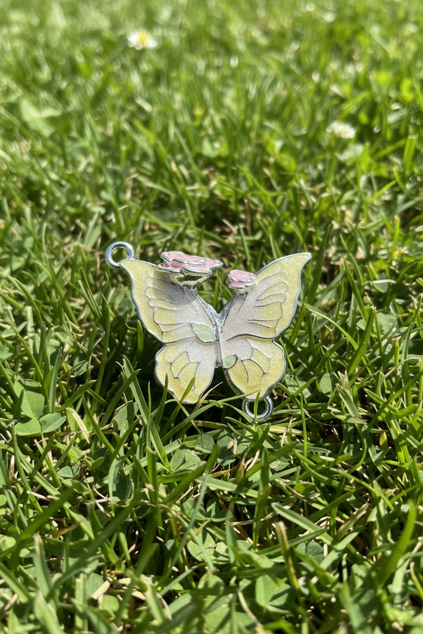 A butterfly-shaped jewellery charm with pale yellow and white enamel wings, silver-tone metal outlines, and small pink flower accents at the top.