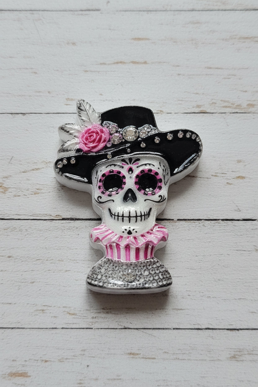 A Day of the Dead sugar skull bead featuring a silver and white skull with pink accents, wearing a black hat with a pink rose and a pink striped dress.