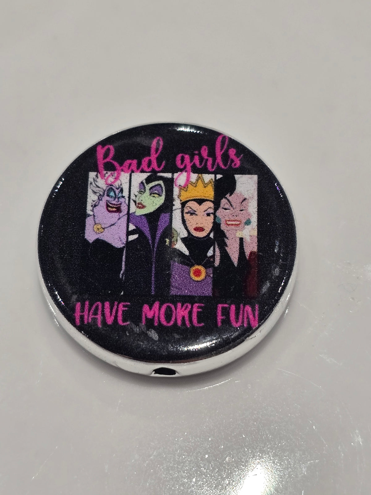 A round acrylic bead featuring a glossy black background with illustrations of four female villains and pink text reading 'Bad girls HAVE MORE FUN'.