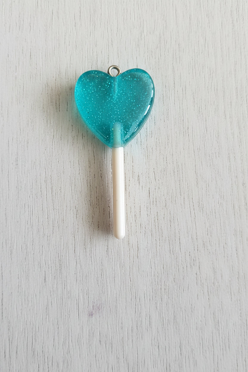 A heart-shaped translucent turquoise lollipop pendant with a white stick and a small metal loop at the top.