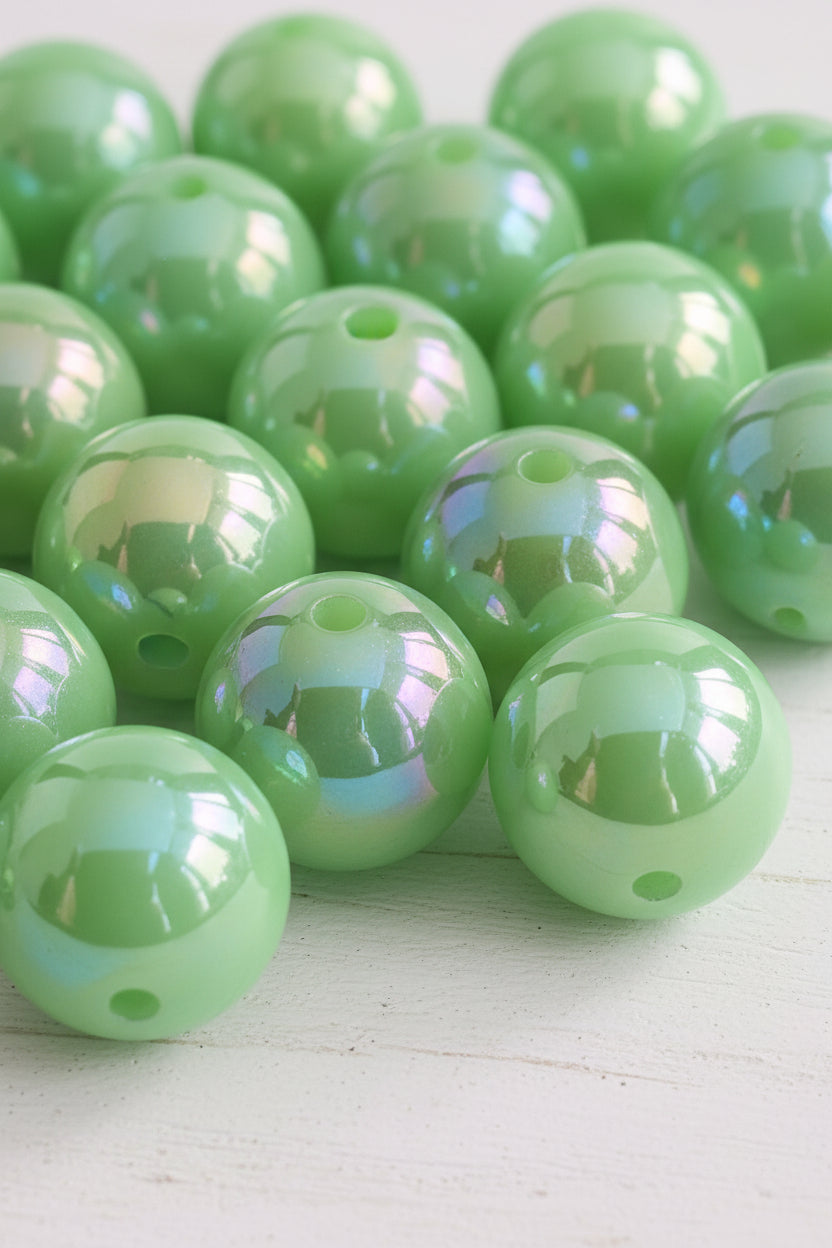 A close-up pile of glossy, light green resin beads with pre-drilled holes, reflecting light and showing a dark background.