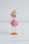 A small duck pendant. The duck has a yellow knit hat, a pink sweater, a white body, and orange feet and beak.