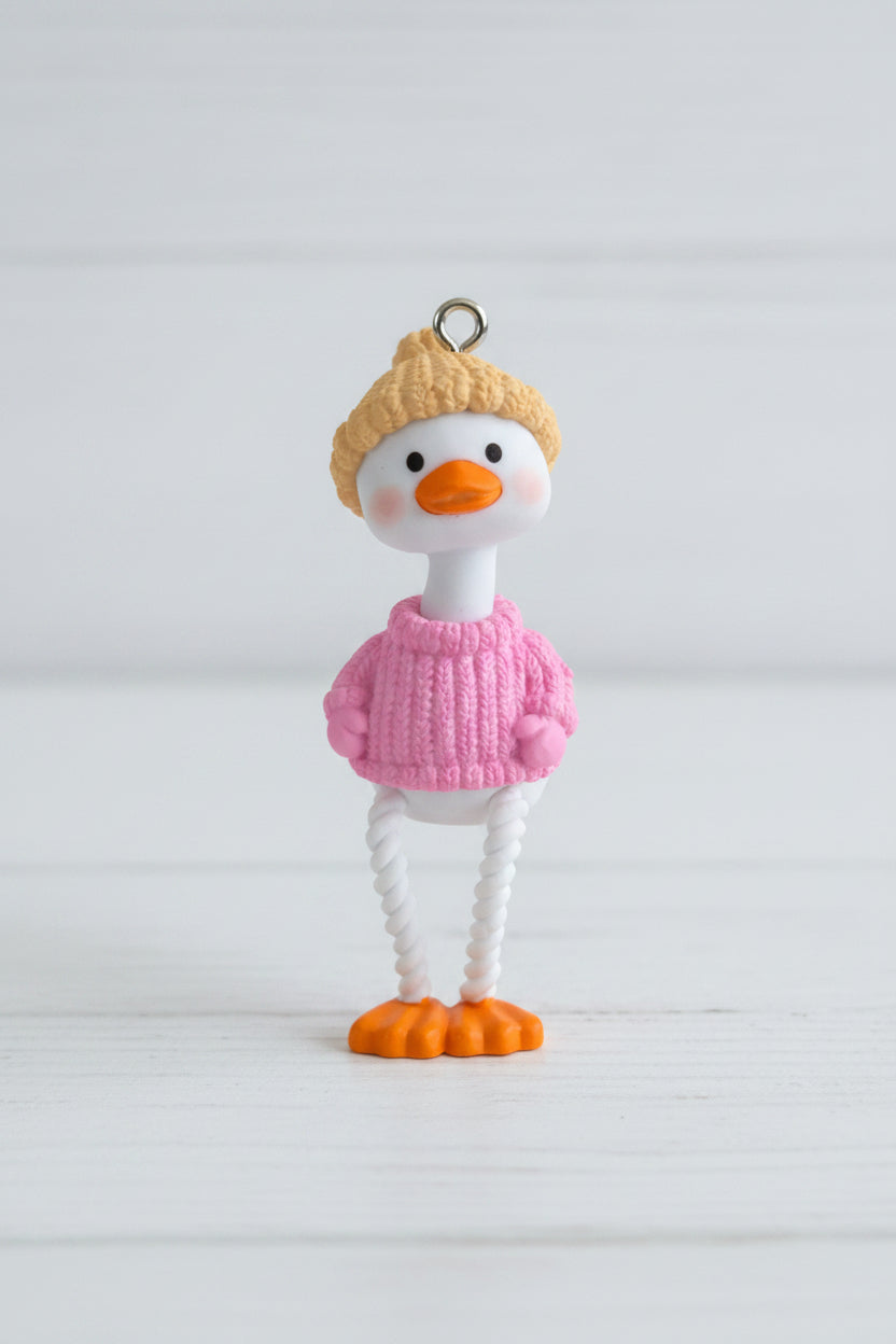 A small duck pendant. The duck has a yellow knit hat, a pink sweater, a white body, and orange feet and beak.