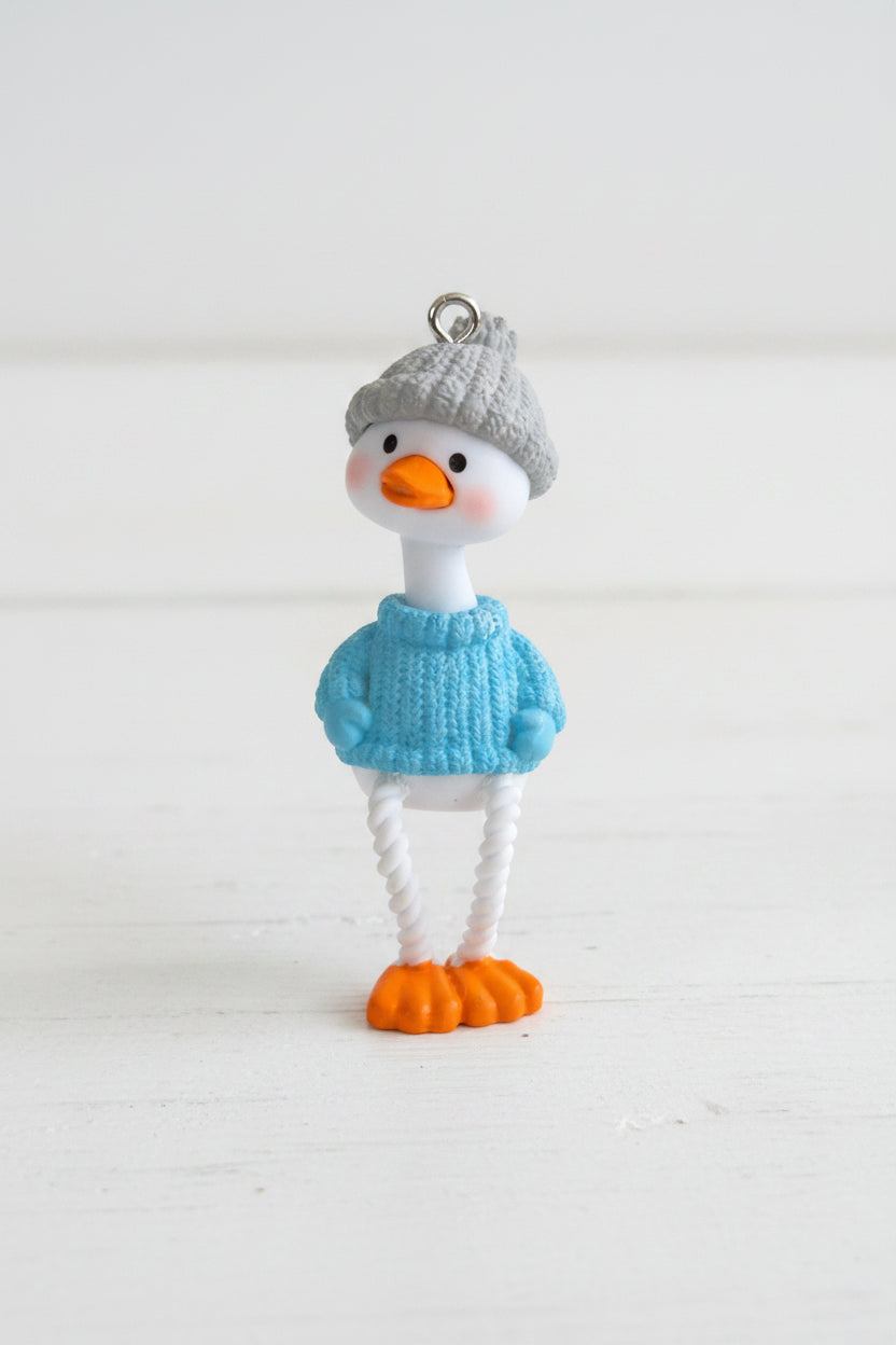 A small duck pendant. The duck has a white body, orange beak and feet, a gray knit hat, and a light blue knit sweater.