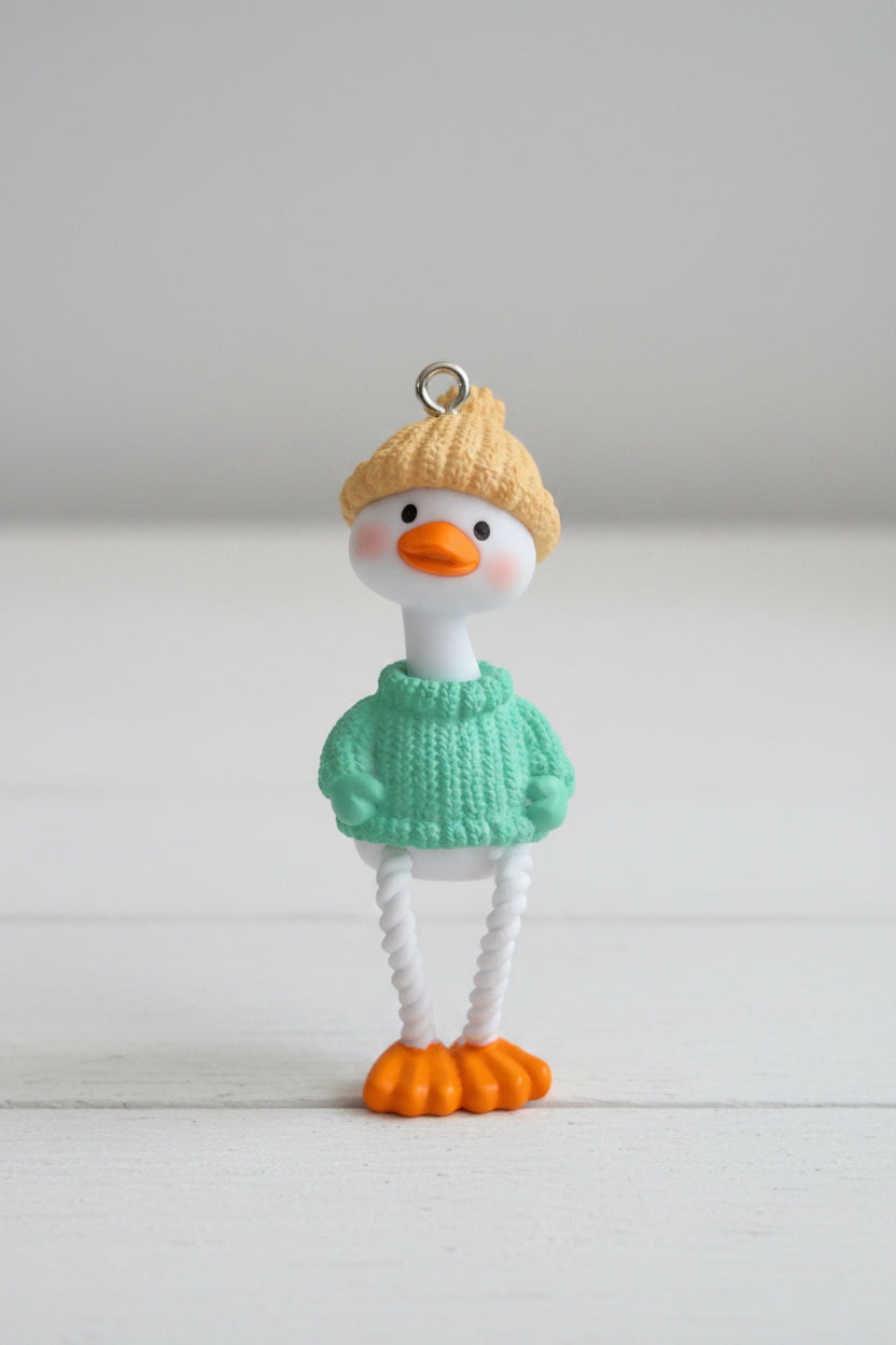 A small duck pendant. The duck has a white body, orange beak and feet, a yellow knit hat, and a mint green knit sweater.