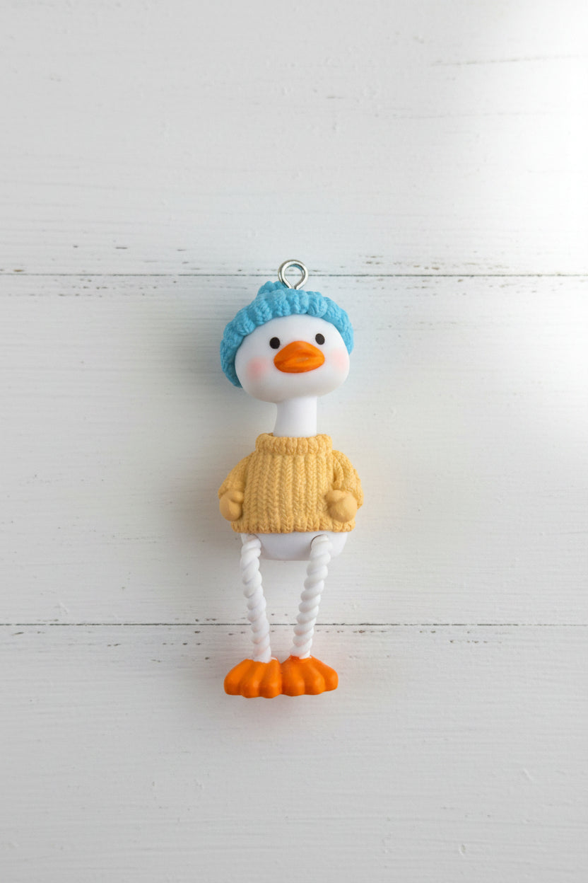 A small duck pendant. The duck has a white body, orange beak and feet, a blue knit hat, and a yellow knit sweater.