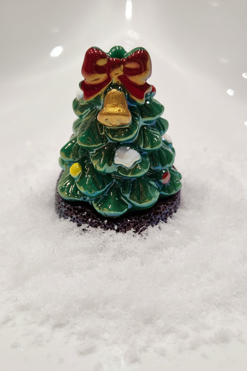 A small Christmas tree bead with green layered branches, a red bow, a gold bell, and a speckled dark base.