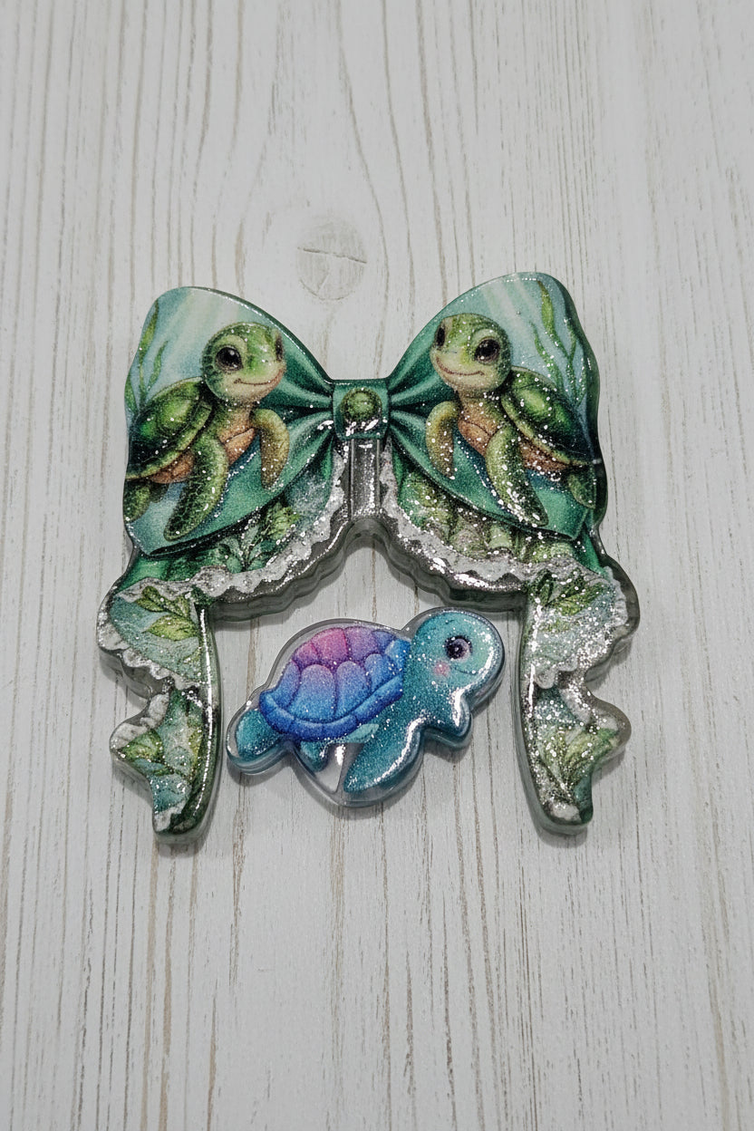 An acrylic bow and bead set featuring illustrated green sea turtles, with a smaller blue and purple turtle charm