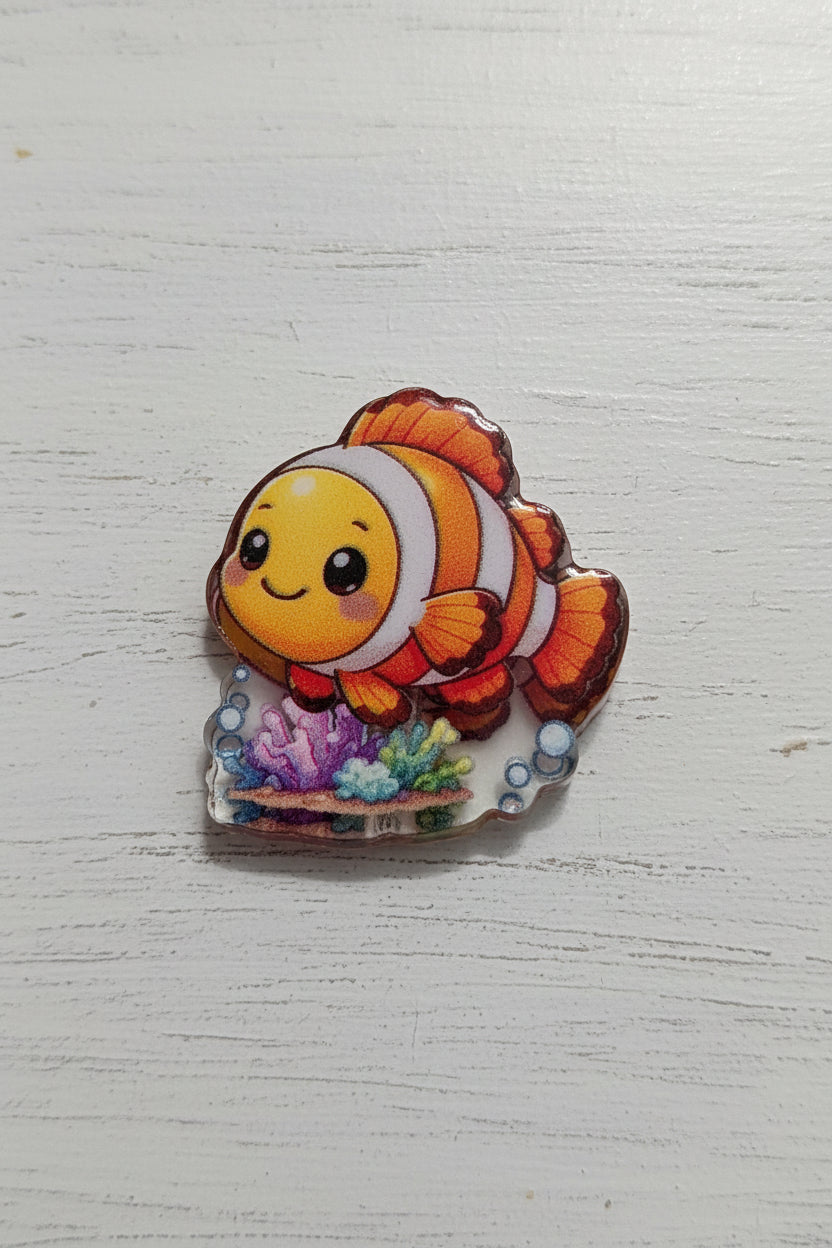 A small fish-shaped bead with a smiling clownfish design in orange, white, and yellow, attached to a clear base with colorful coral details.