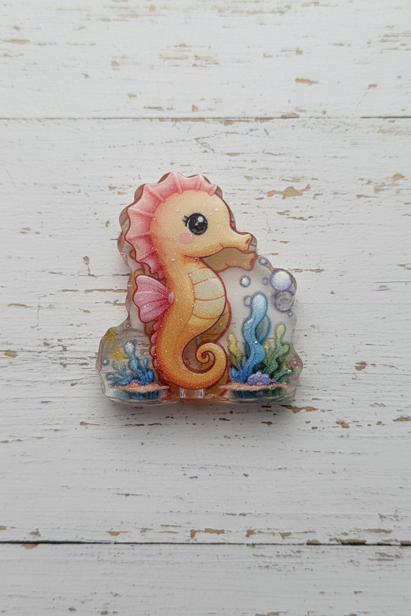 A small decorative seahorse bead with a pink and orange body, set in a clear acrylic base with blue and green underwater details.
