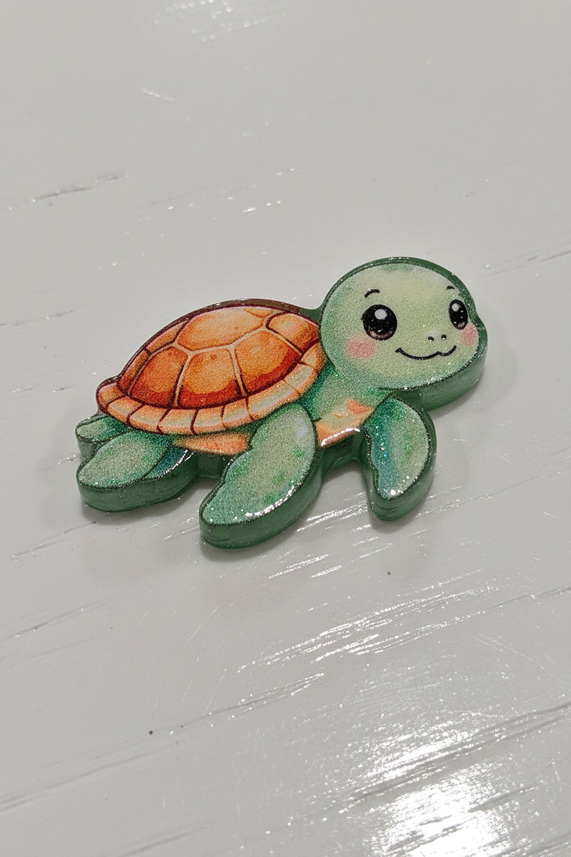 A small painted turtle bead with a green head and legs, a brown shell, and pink cheeks.