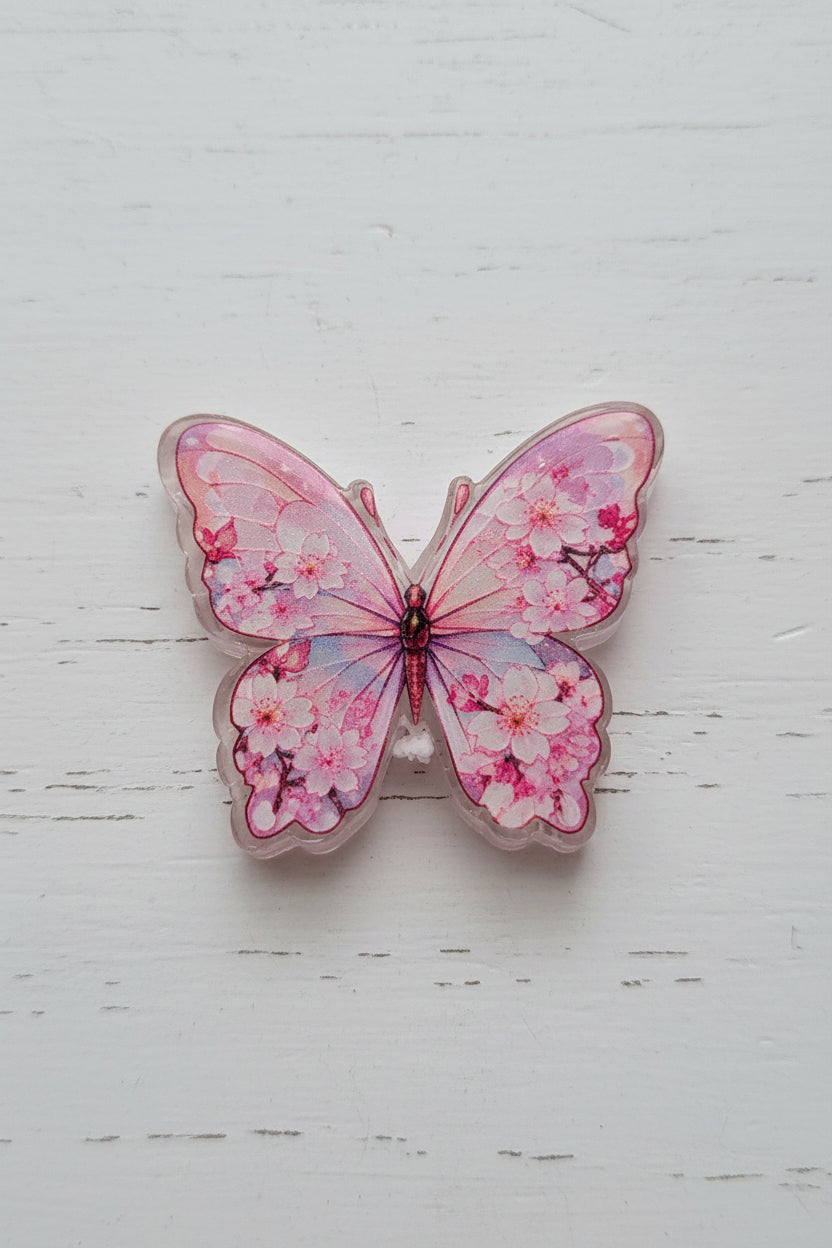A butterfly-shaped resin charm or pendant with pink cherry blossom patterns and glitter.