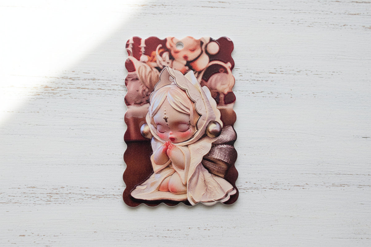 A rectangular scalloped tag featuring a stylized praying doll illustration in muted pink, beige, and burgundy tones
