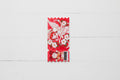 A red tag with a scalloped bottom edge, featuring anime-style artwork of a white-haired character, cherub wings, cherry blossoms, and a small cartoon figure near the bottom. A barcode sticker is attached on the lower right.