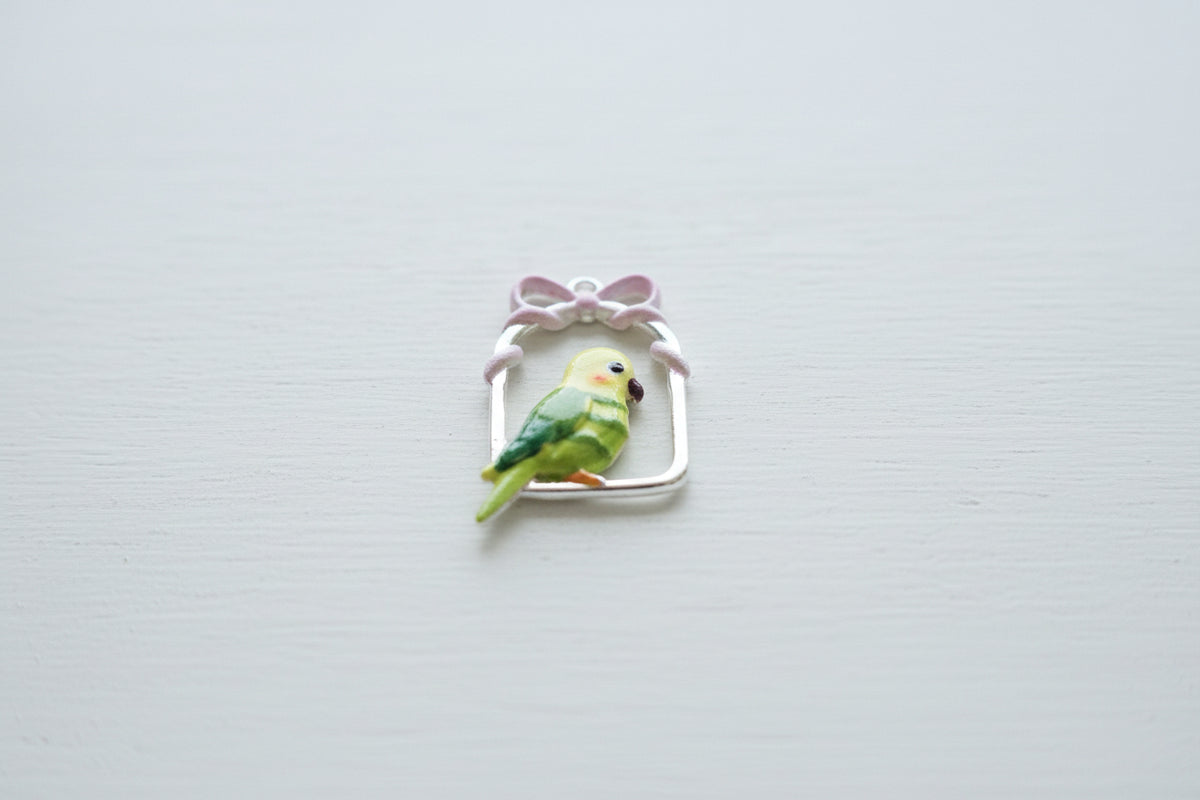 A small enamel bird charm shaped like a green and yellow parrot perched inside a white arched frame with a pink bow on top.