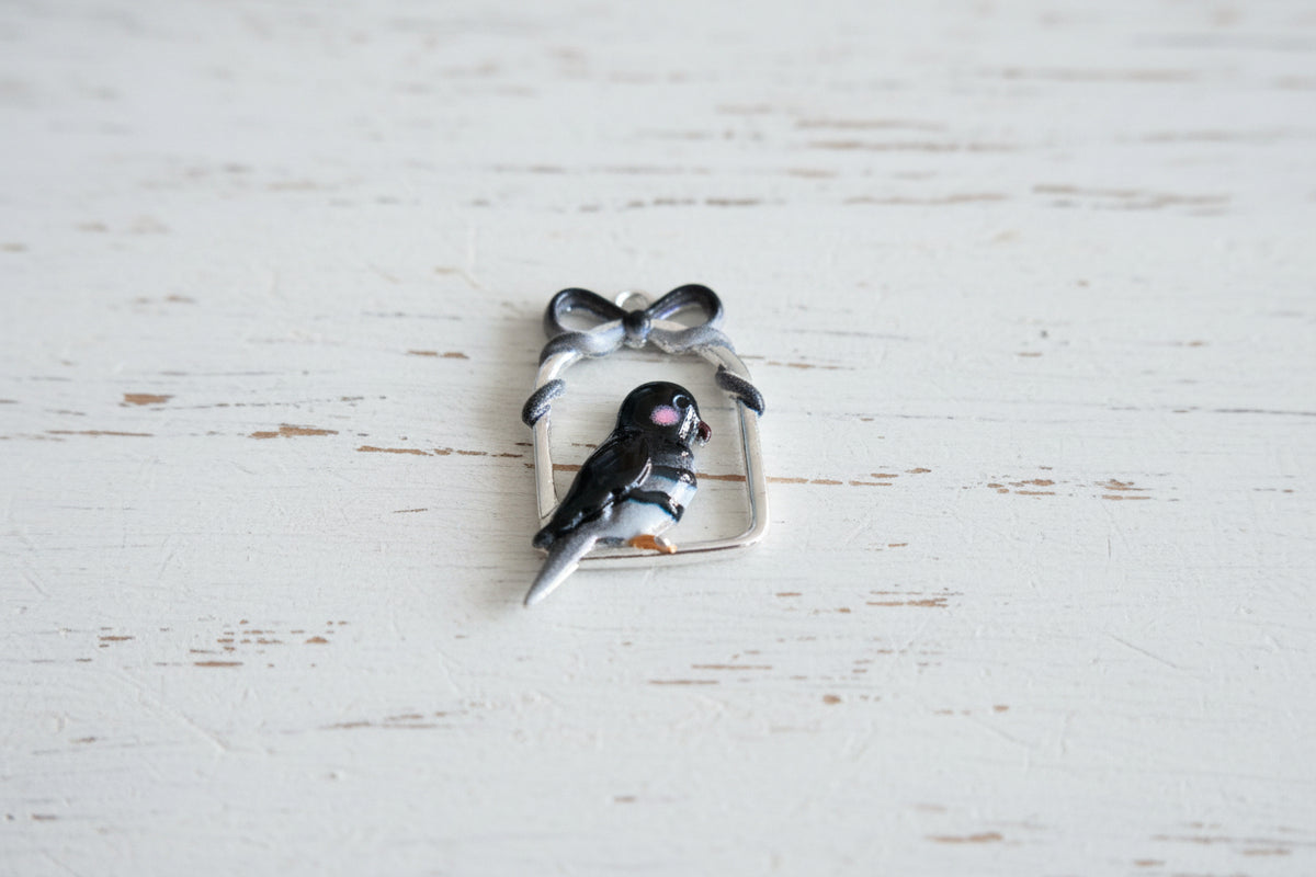 A small bird charm with a black and white body, pink cheek, and orange beak, framed by a silver bow and rectangular border.