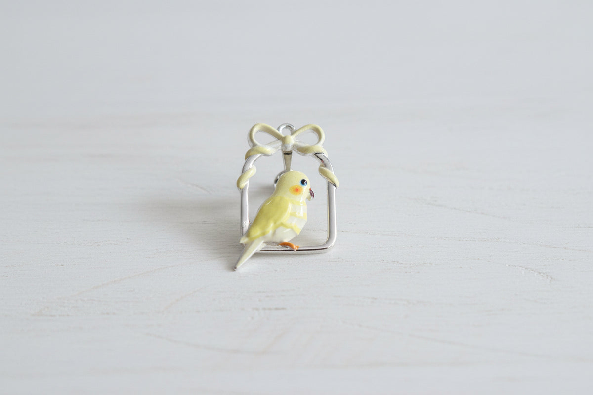 A small yellow bird charm with orange cheek patches and a black eye, perched inside a twisted cream-colored metal frame with a bow at the top.