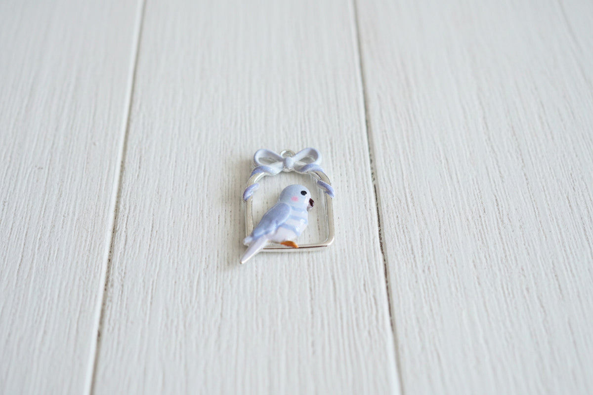 A small bird-shaped jewelry charm or pendant with a pale purple  body, pink cheek, set inside a white bow-shaped frame 