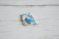 A small enamel bird charm in light blue and white, perched inside a silver-toned rectangular frame with a bow at the top.