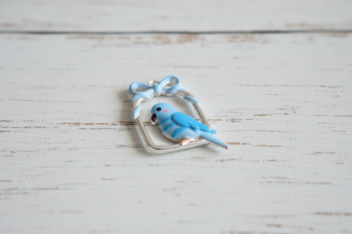 A small enamel bird charm in light blue and white, perched inside a silver-toned rectangular frame with a bow at the top.