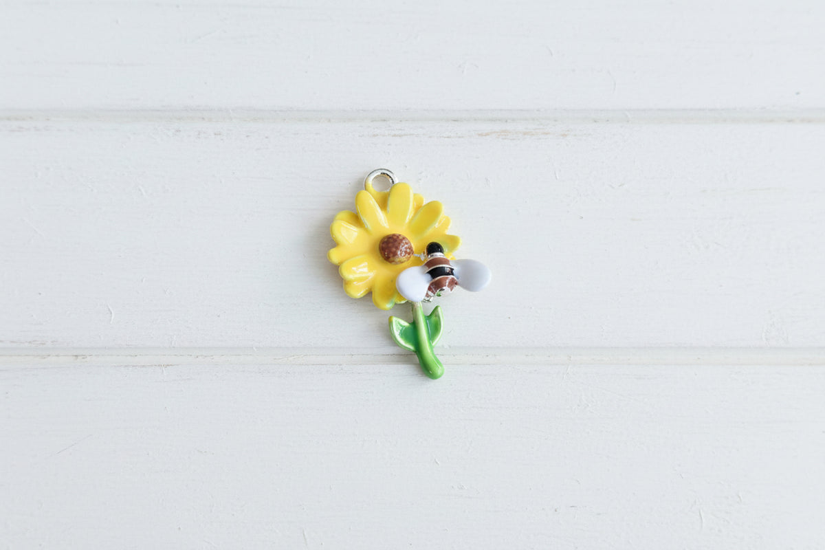 A small enamel charm shaped like a yellow sunflower with a bee on the stem.