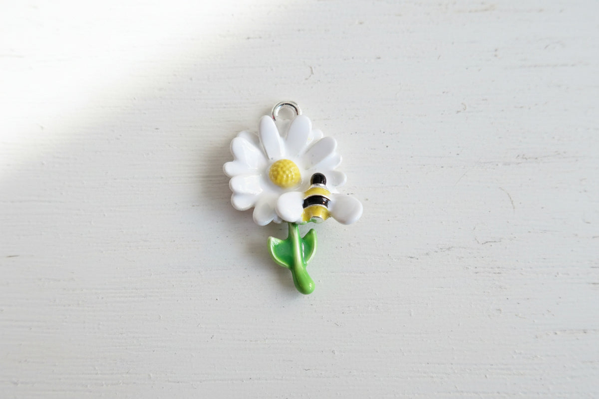 A small enamel charm shaped like a white daisy with a yellow center, green stem, and a yellow-and-black bumblebee on it.