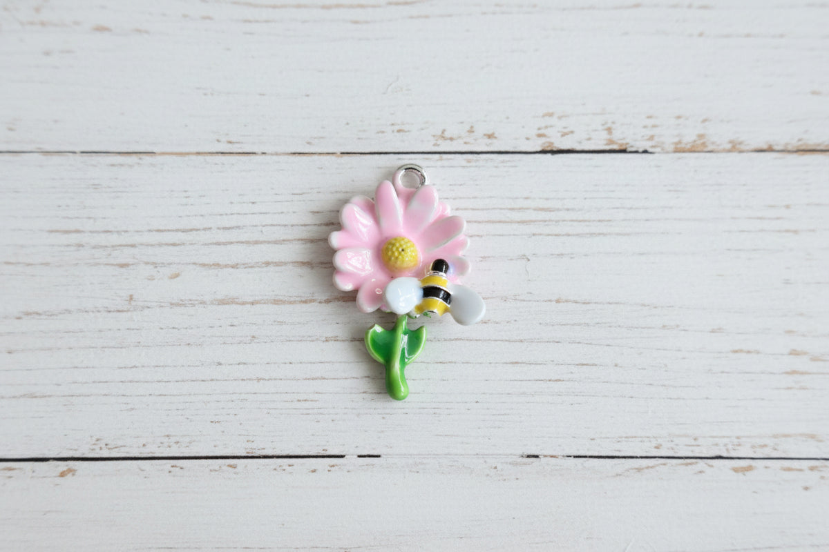 A small enamel charm shaped like a pink flower with a yellow center, green stem and leaves, and a yellow-and-black bumblebee attached.