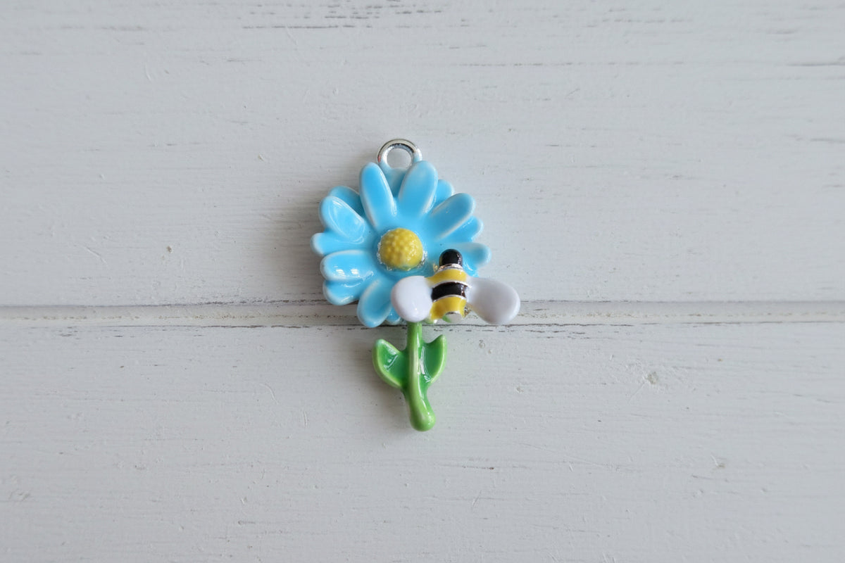 A small enamel charm featuring a light blue flower with a yellow center, green stem and leaves, and a yellow-and-black bumblebee perched on the side.