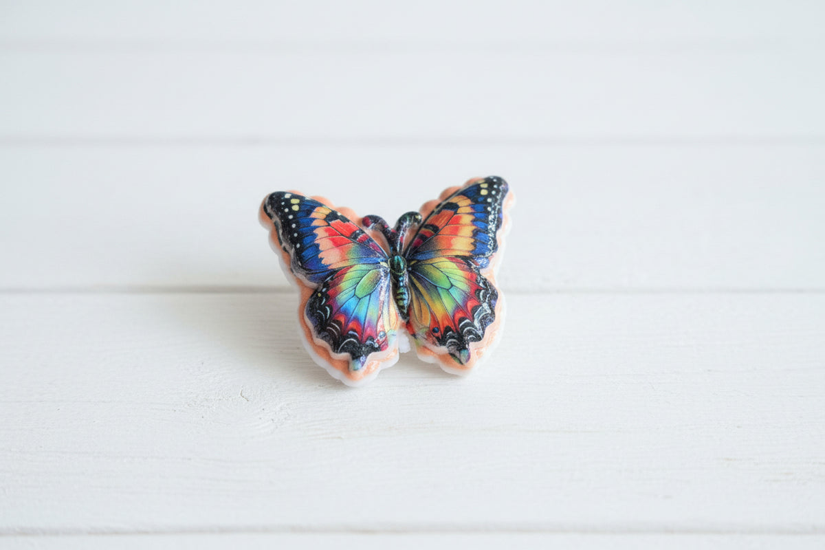 A butterfly-shaped acrylic bead with multicolored wings in blue, red, yellow, green, and orange, outlined in black and set against a peach-pink background.