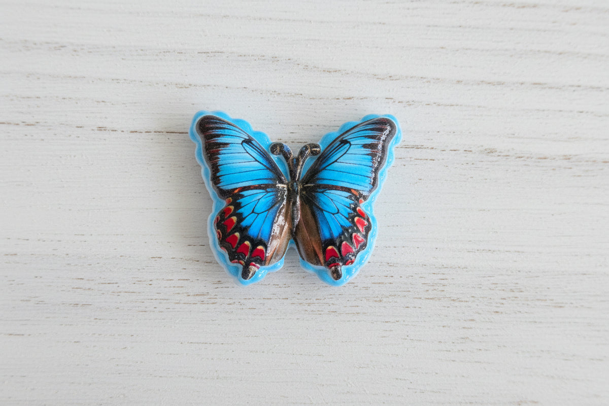 A butterfly-shaped acrylic bead with vibrant blue wings, black veining, and red and brown accents