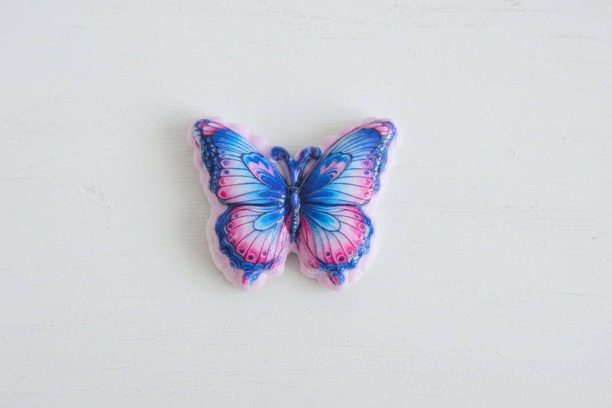 A butterfly-shaped acrylic bead with blue and pink marbled wings and a scalloped pink edge.