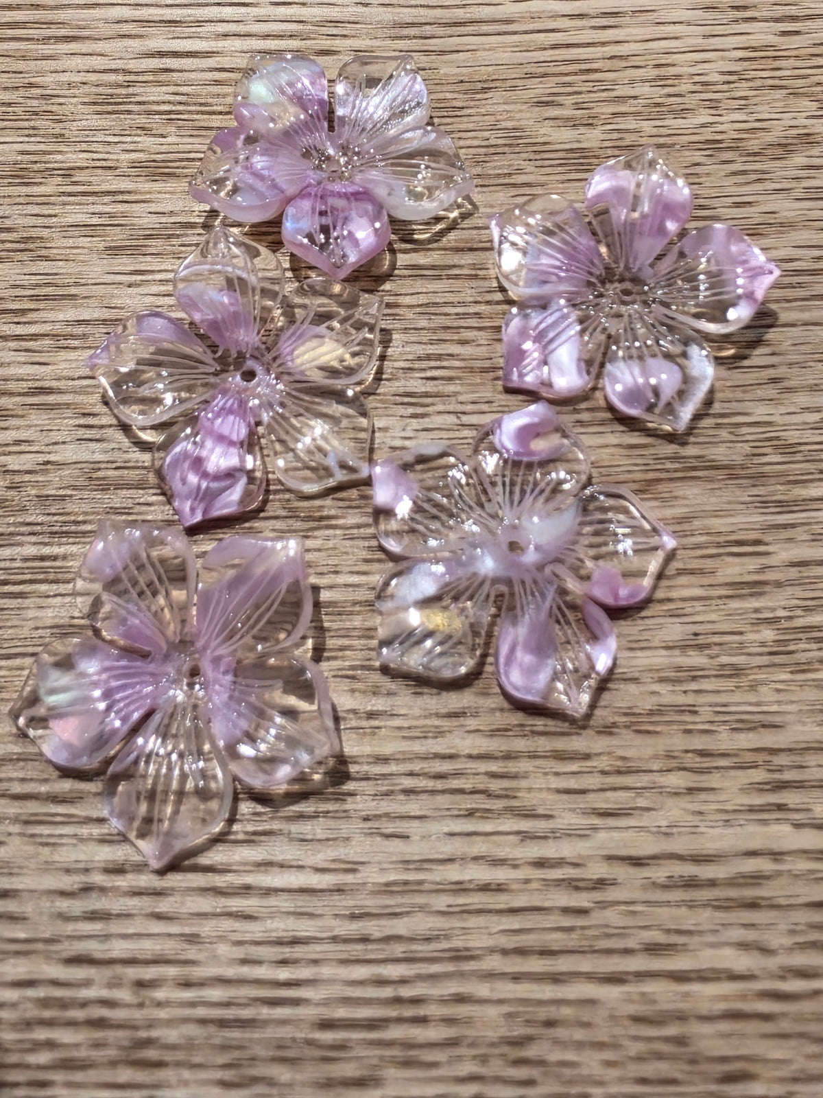 Lavender and pink cherry blossom flower charms with iridescent highlights, arranged on a light brown wood grain surface.