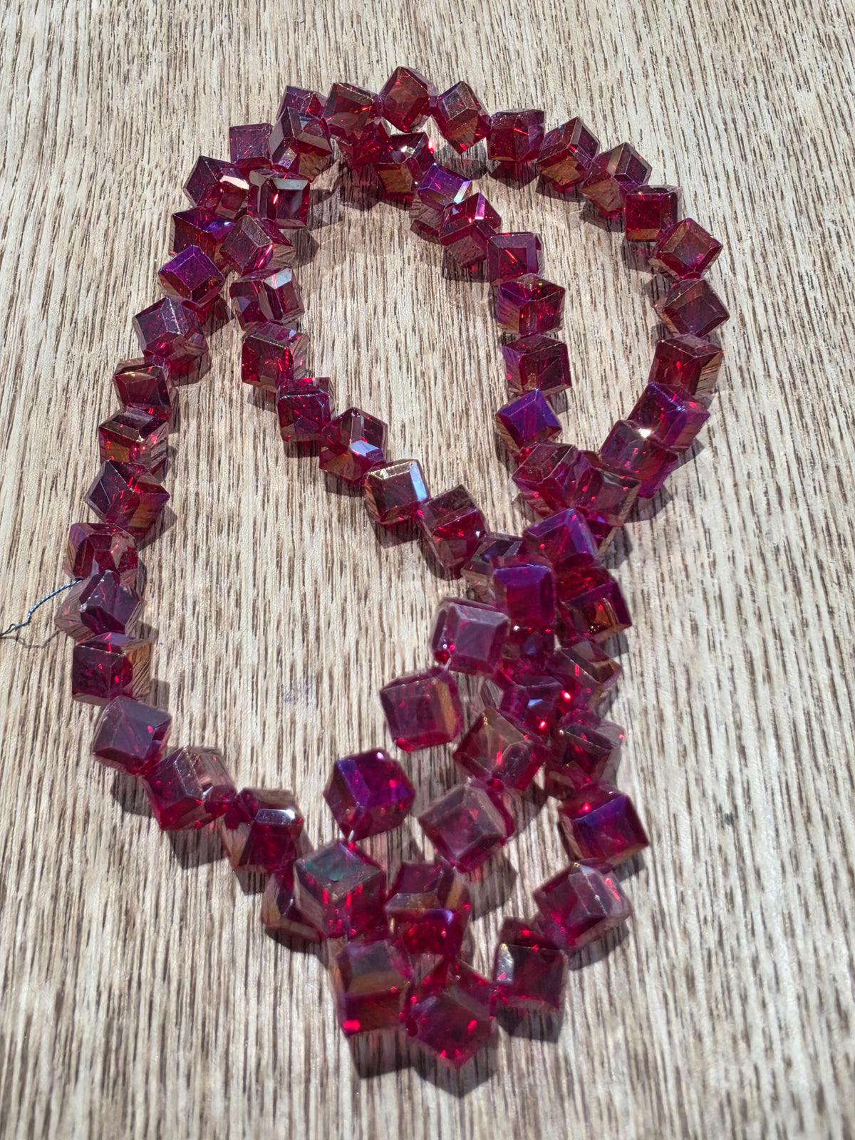 A strand of  cube glass beads in a red color, arranged in a loop on a light-colored wooden surface.