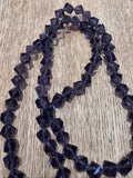 A strand of faceted dark purple glass beads,  arranged in loops on a light brown wood grain surface.