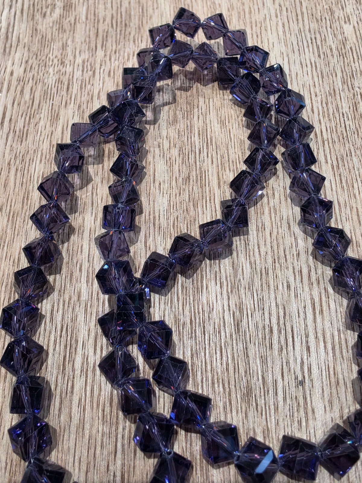 A strand of faceted dark purple glass beads,  arranged in loops on a light brown wood grain surface.