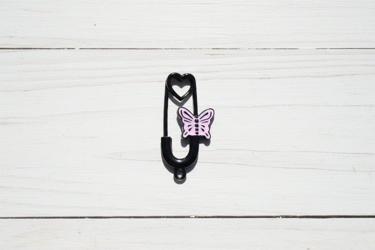 A black safety pin-shaped charm with a small lavender butterfly and heart design, placed on a light wood grain surface.