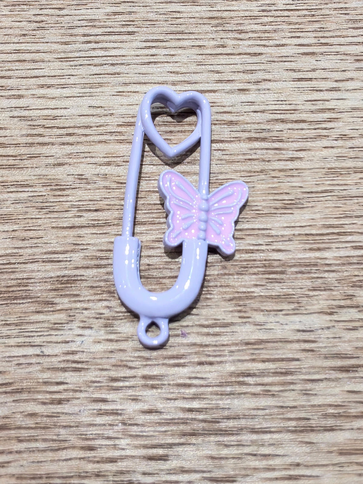 A small safety pin-shaped charm in light lavender purple with a pink butterfly accent, placed on a light brown wood-grain surface.