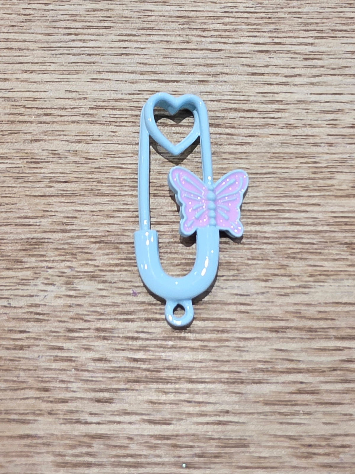 A light blue safety pin charm with a heart-shaped top loop and a small pink butterfly accent on the side, resting on a wooden surface.