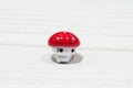 A small mushroom-shaped bead with a red cap and white body, resting on a textured wooden surface.