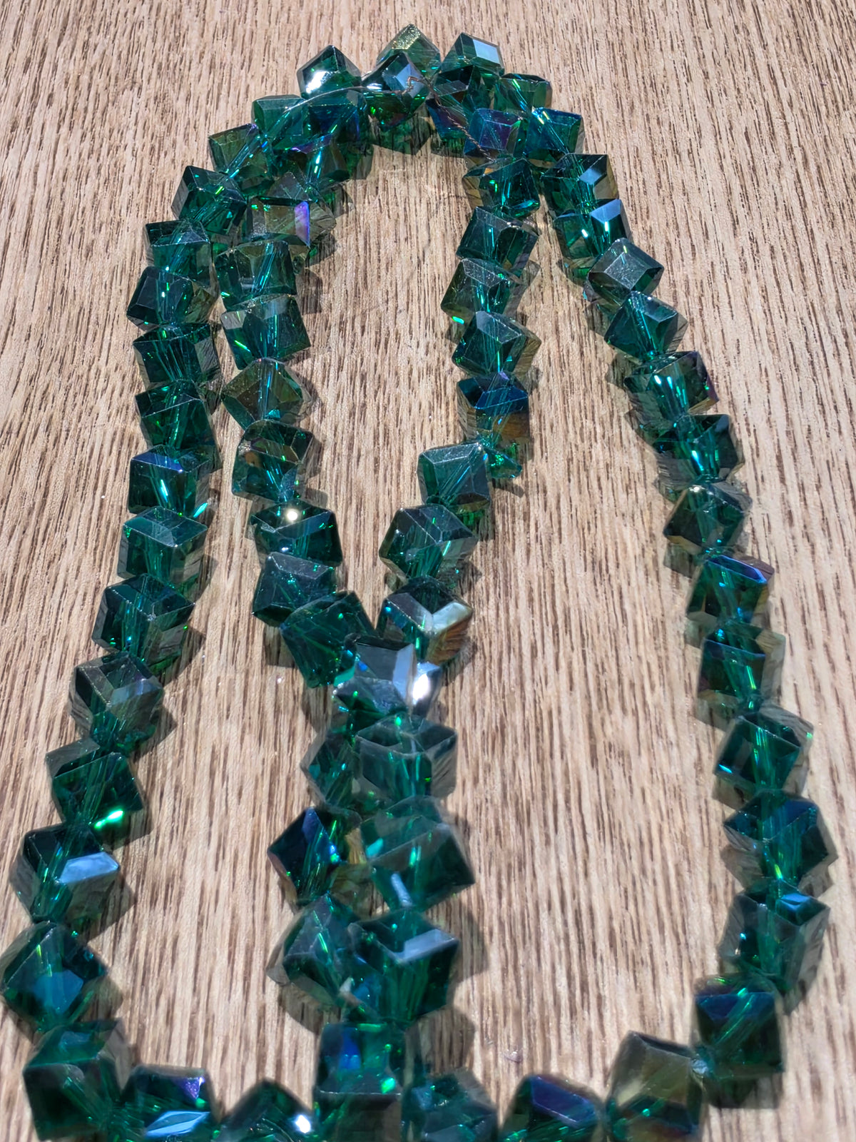 A strand of glass crystal beads in a deep green color, arranged in loops on a light wood grain surface.