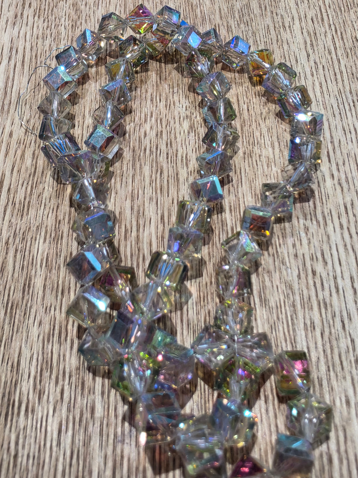 A strand of glass cube beads with an iridescent, rainbow-reflective coating, arranged on a light brown wood grain surface.