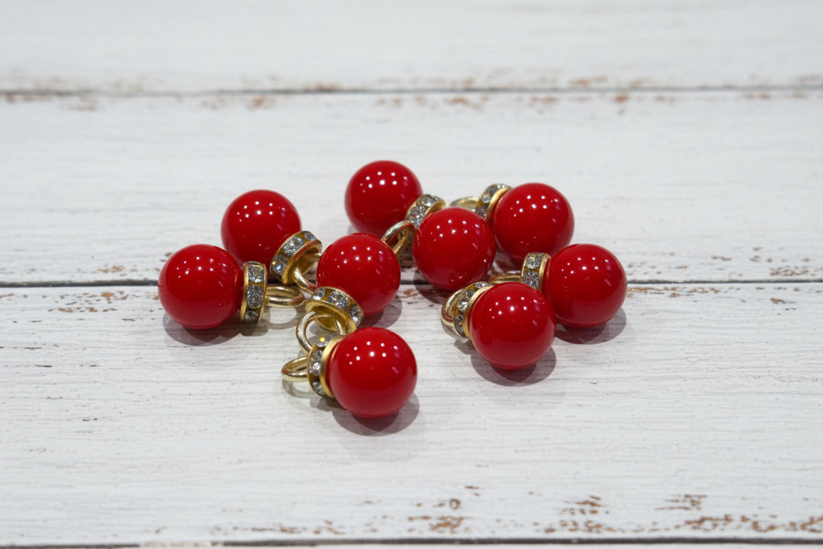 red pearl-like pendant beads with gold-toned metal loops and small clear rhinestone accents, arranged on a light wood surface.