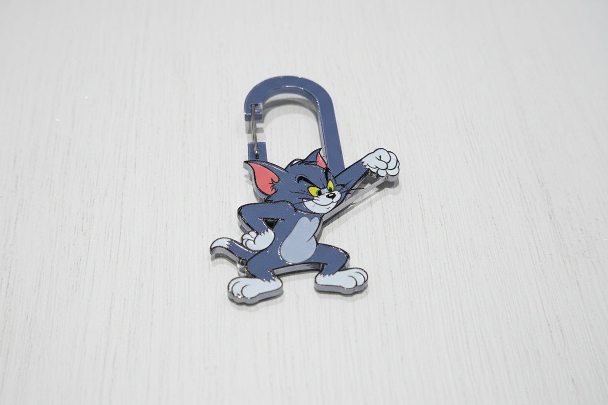 A cute cat keyring featuring the cartoon cat Tom in a dancing pose attached to a silver metal carabiner, shown on a wooden surface.