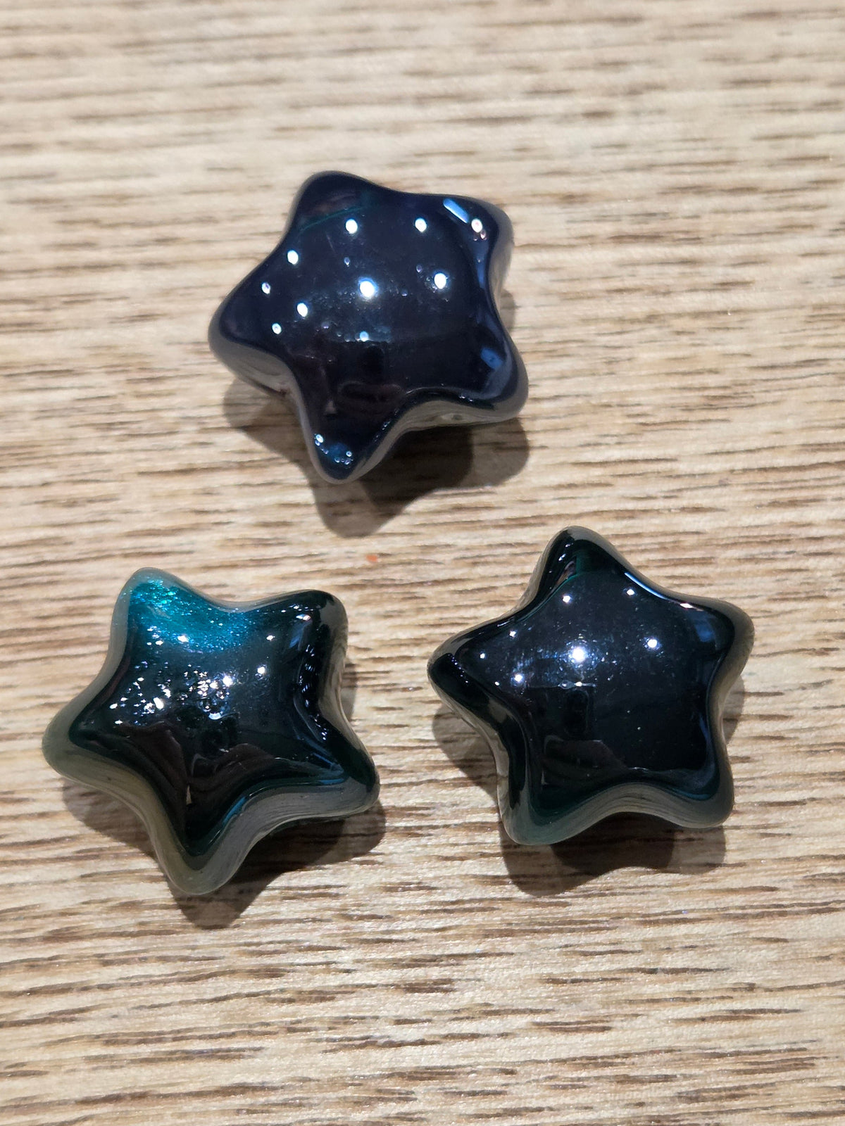 Three glossy star-shaped beads in dark blue and teal colors with white speckles, arranged on a light wood grain surface.